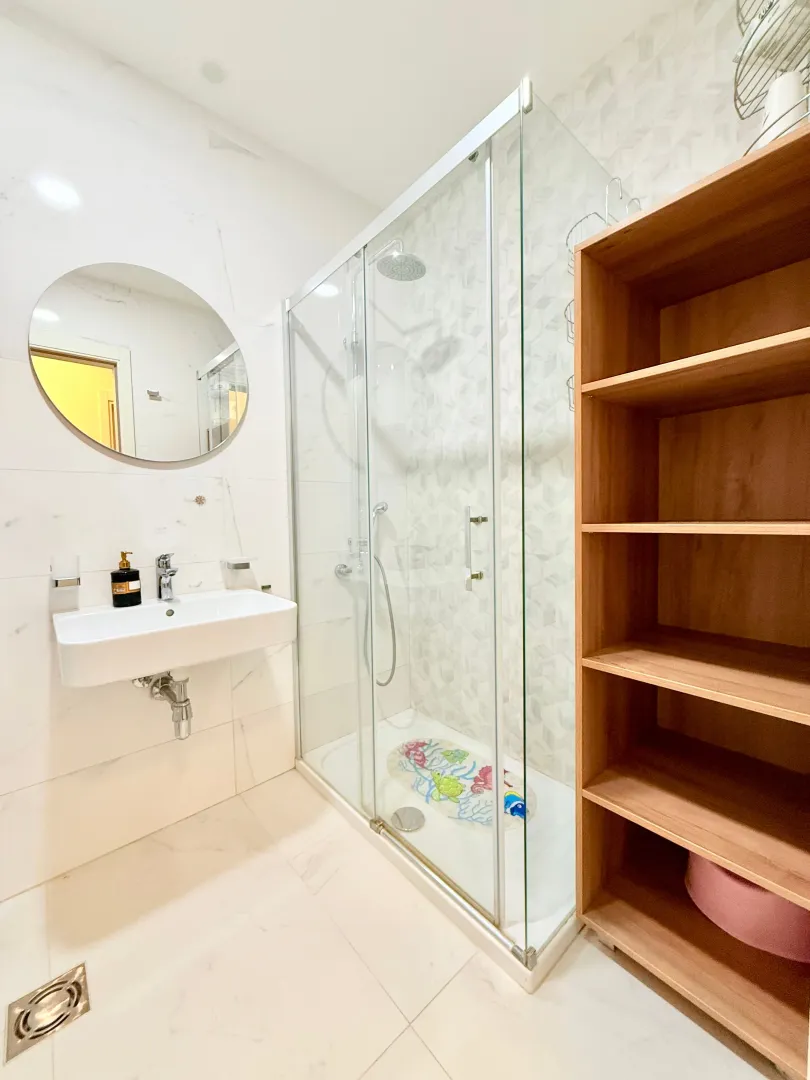 Bright equipped bathroom with a shower cabin, sink, and wooden shelving unit.