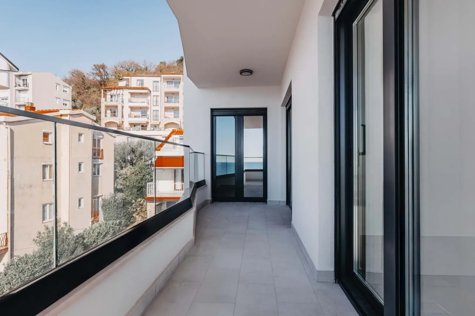 Prostrani balkon sa staklenom ogradom i pogledom na okolne zgrade i more.