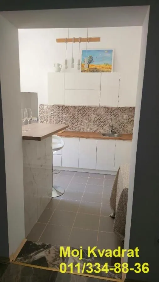 Pogled iz prolaza na manju kuhinju sa belim elementima i šankom.