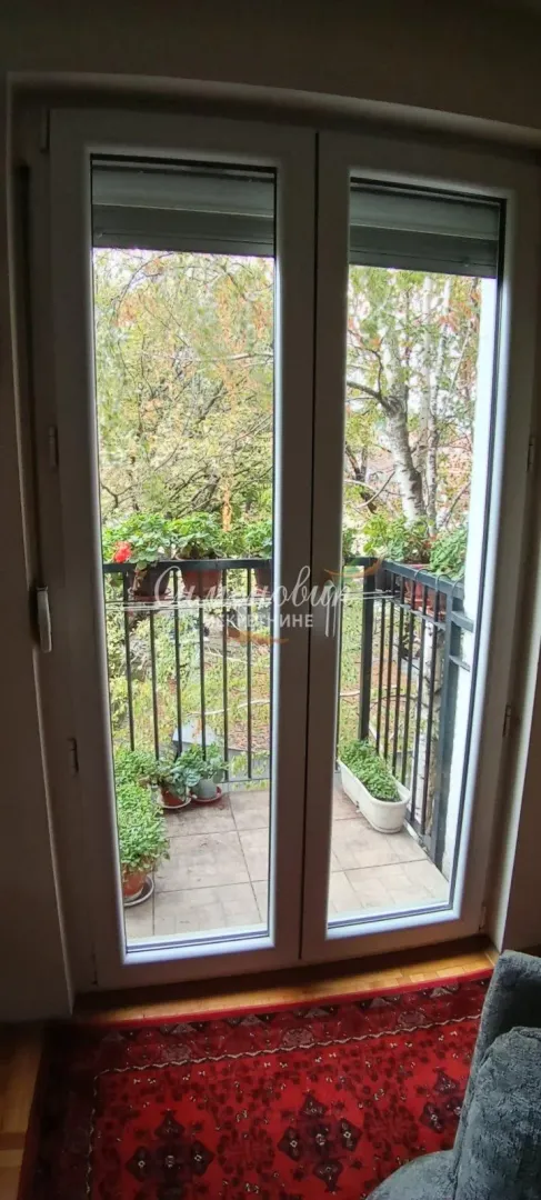 Pogled kroz balkonska vrata na uređeni balkon sa zelenilom.