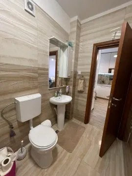 Kupatilo obloženo bež pločicama sa šoljom, umivaonikom i ogledalom. - image 4
