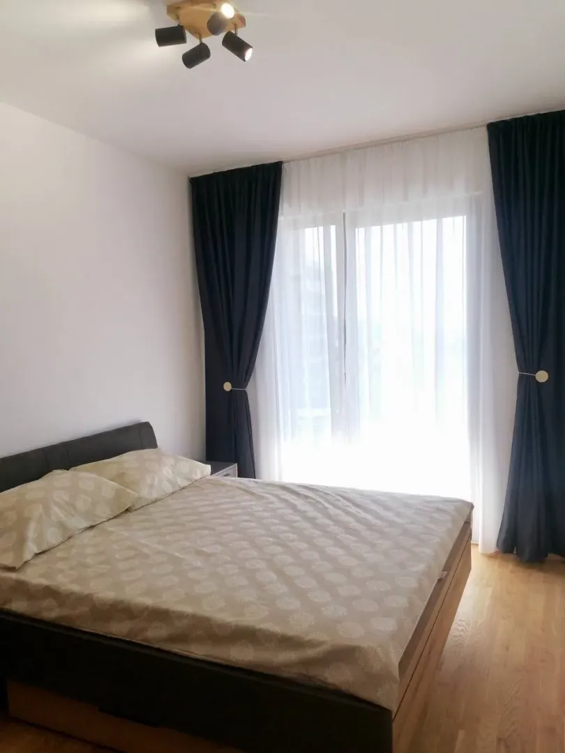 Spavaća soba sa bračnim krevetom, prozorom sa tamnim zavesama i plafonskim osvjetljenjem.