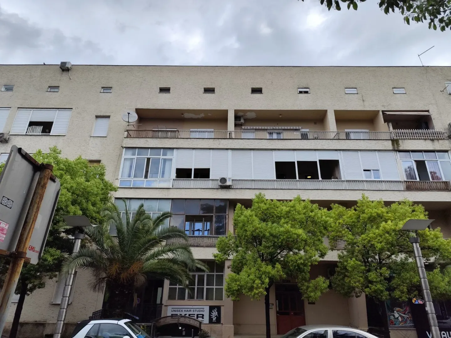 Pogled na fasadu višemilnospratne stambene zgrade sa balkonom i zelenilom.