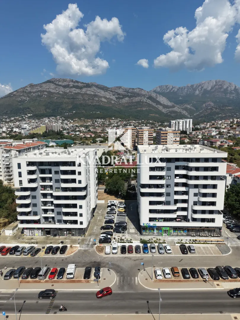Pogled iz vazduha na moderne stambene zgrade sa parkingom i planinama u pozadini.