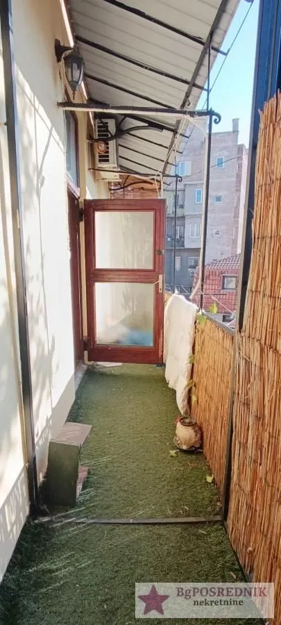 Uski natkriveni balkon sa veštačkom travom na podu i pogledom na okolne zgrade.
