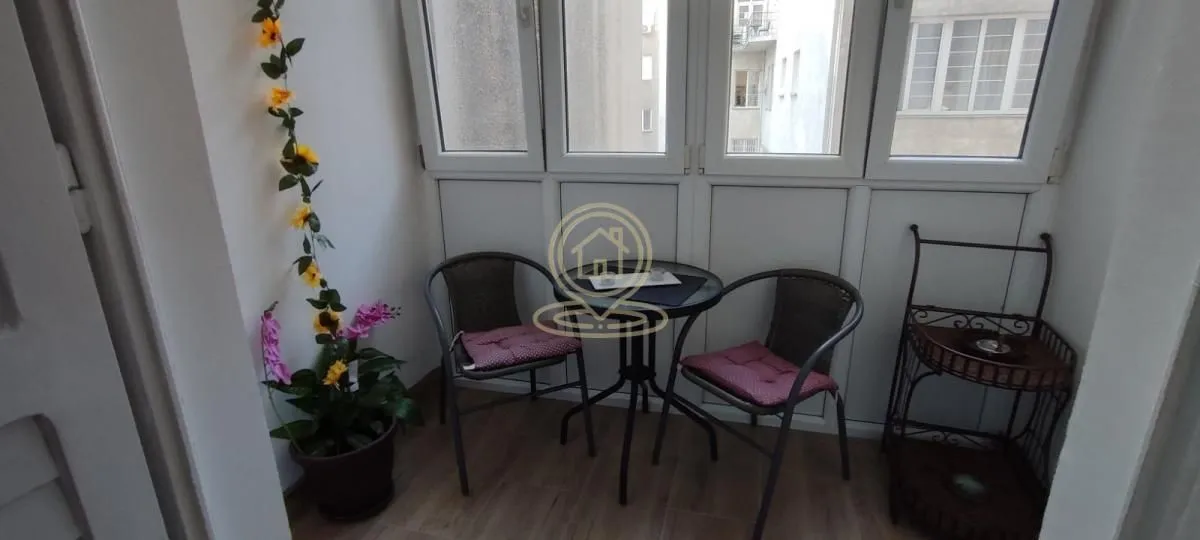 Zastakljeni balkon opremljen stolom, dve stolice i dekorativnim biljkama.