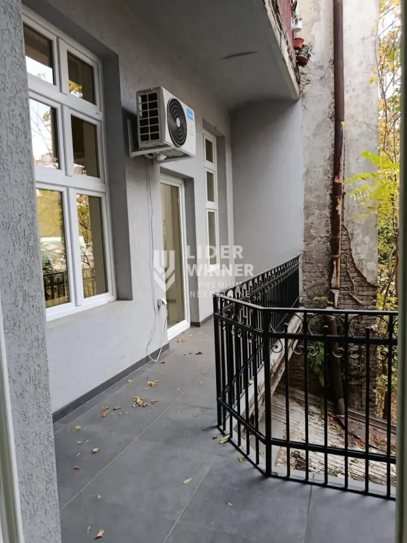 Dugački balkon sa metalnom ogradom i spoljnom jedinicom klima uređaja na zidu.