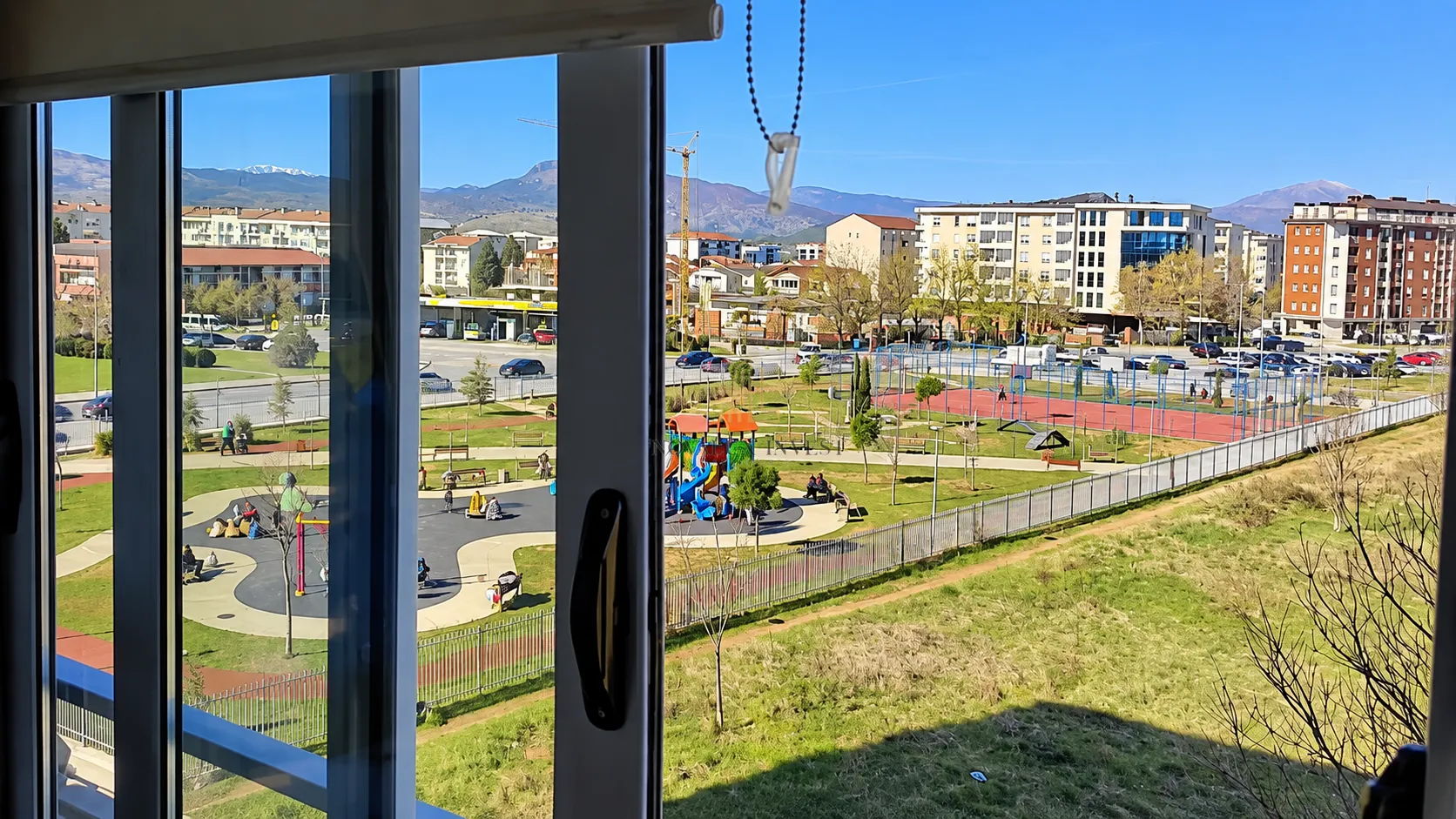 Pogled iz prozora na park, igralište i okolne zgrade sa planinama u pozadini.