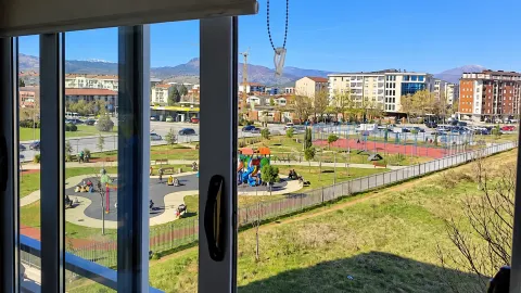 Pogled iz prozora na park, igralište i okolne zgrade sa planinama u pozadini. - image 6