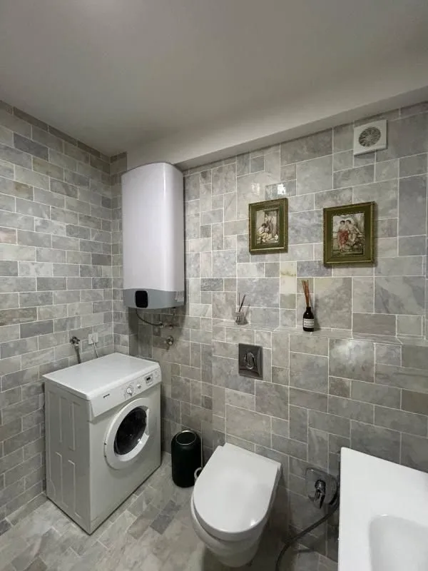 Kupatilo obloženo sivim pločicama sa toaletom, mašinom za veš i bojlerom.