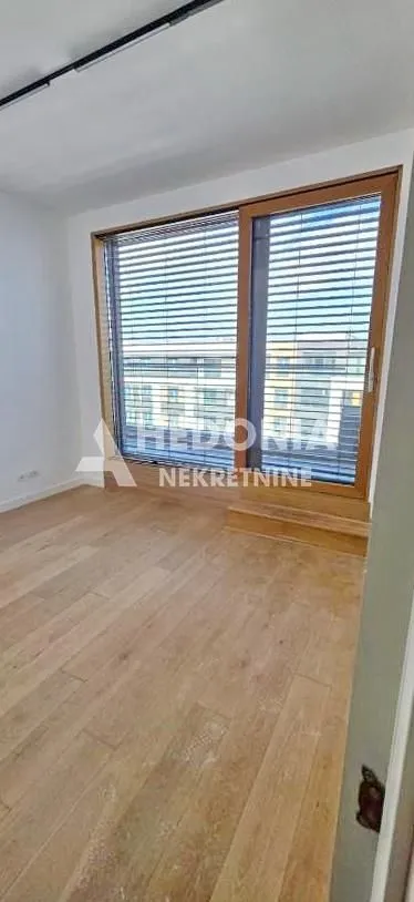 Prazna prostorija sa drvenim podom i velikim balkonskim vratima sa roletnama.