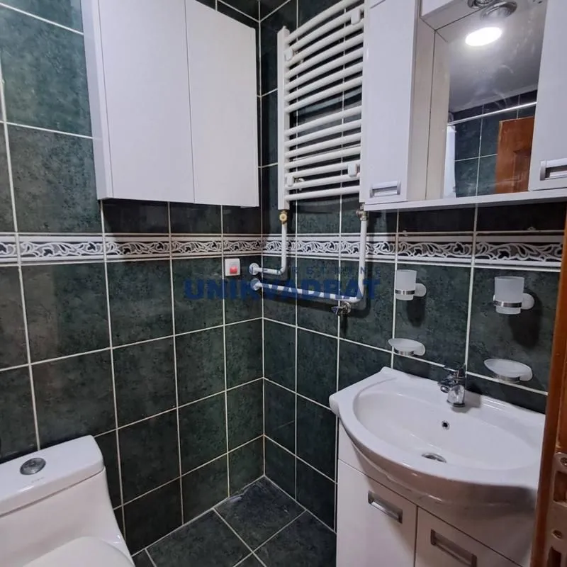 Manje kupatilo obloženo tamnim pločicama sa umivaonikom, WC šoljom i zidnim ormarićem.