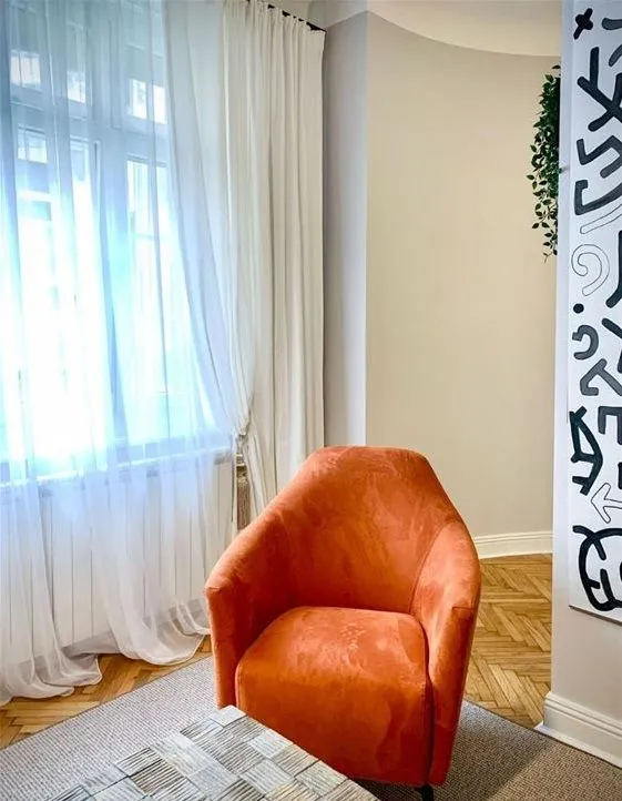 Dnevna soba sa narandžastom foteljom i velikim prozorom sa belim zavesama.