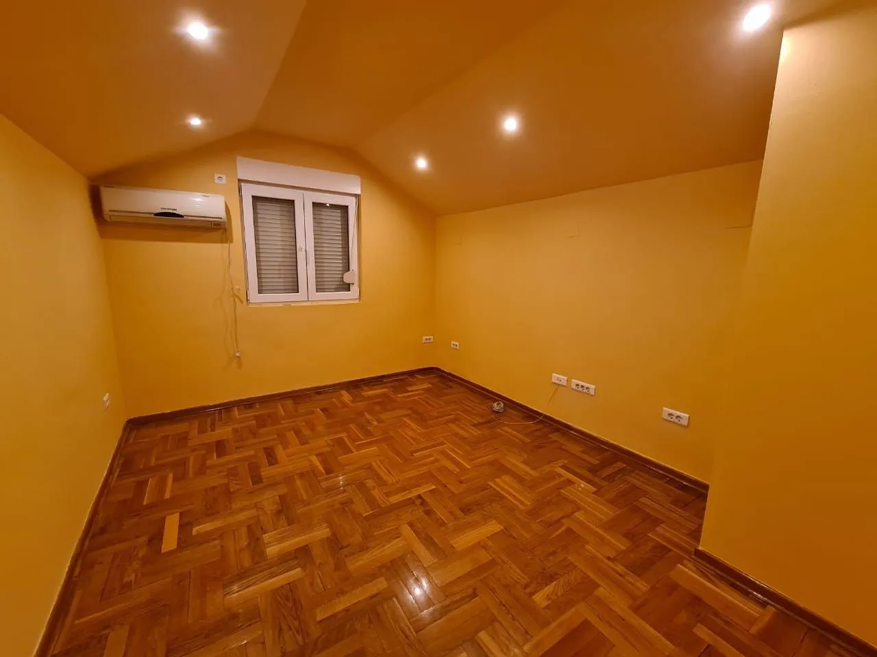Prazna soba sa žutim zidovima, parketom i klima uređajem.