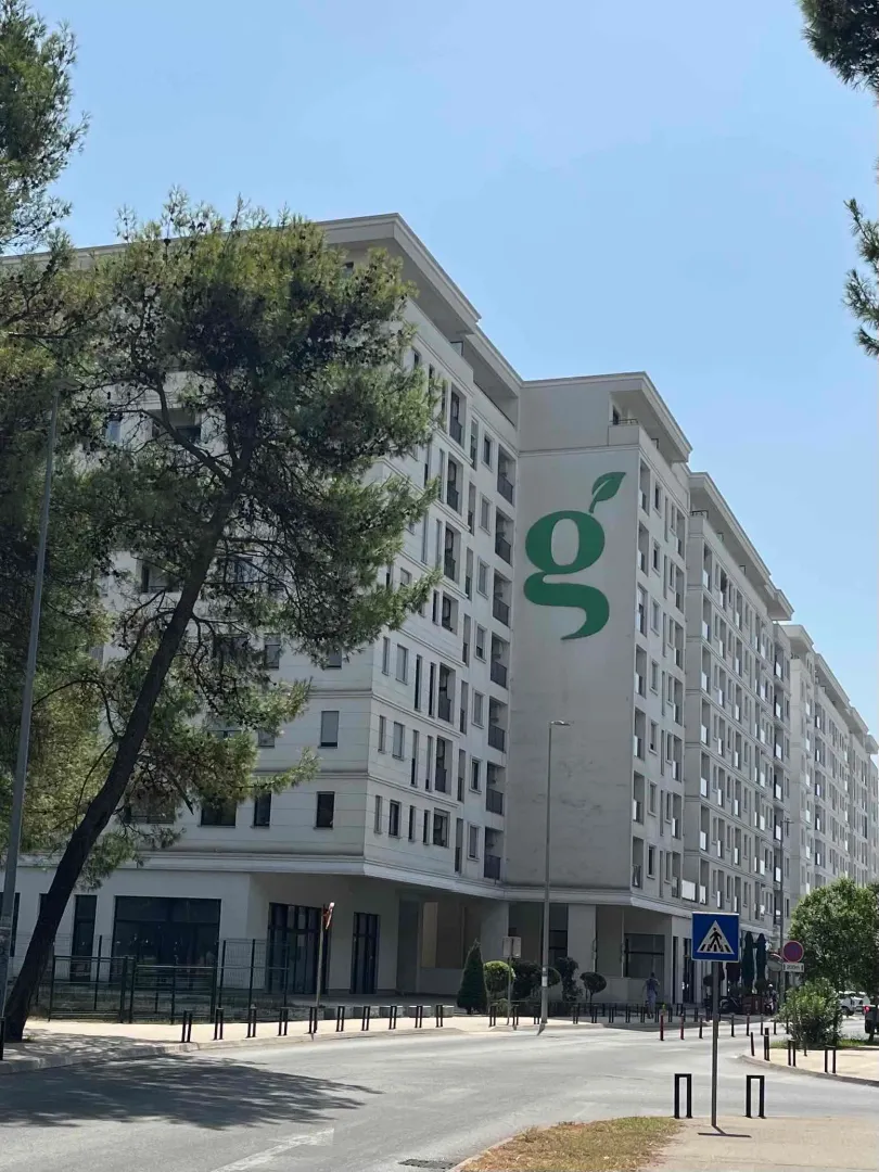 Pogled na spoljašnjost moderne stambene zgrade sa zelenom površinom i ulicom u prvom planu.