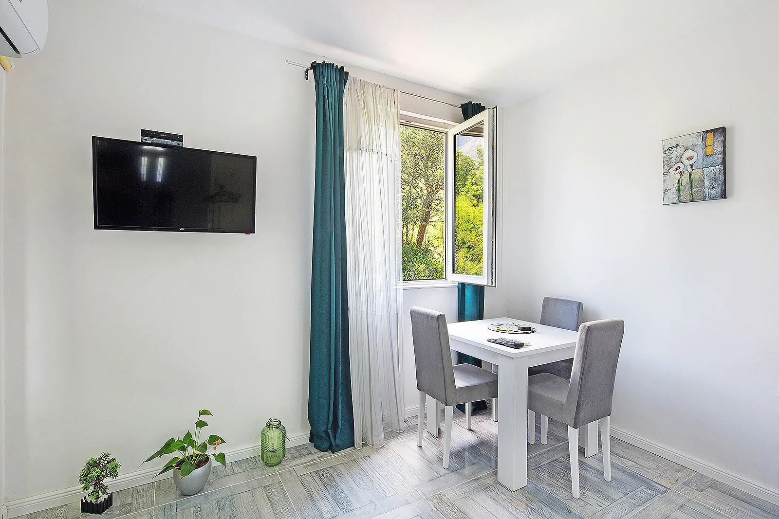 Living room with a dining table, wall-mounted TV, and a window overlooking greenery.
