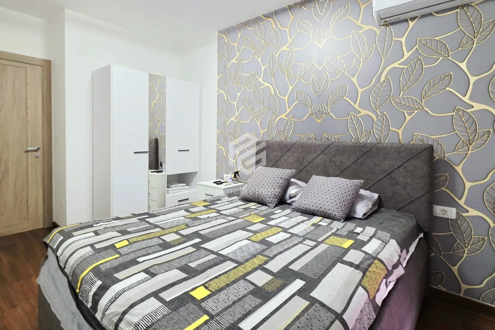 Furnished bedroom with a double bed, white wardrobe, and patterned wallpaper.