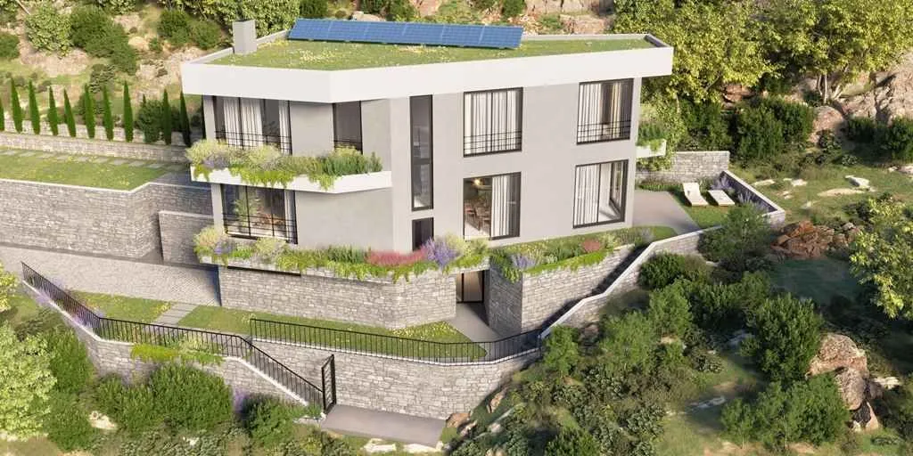 Moderna troetažna kuća na padini sa zelenim krovom i uređenim dvorištem.