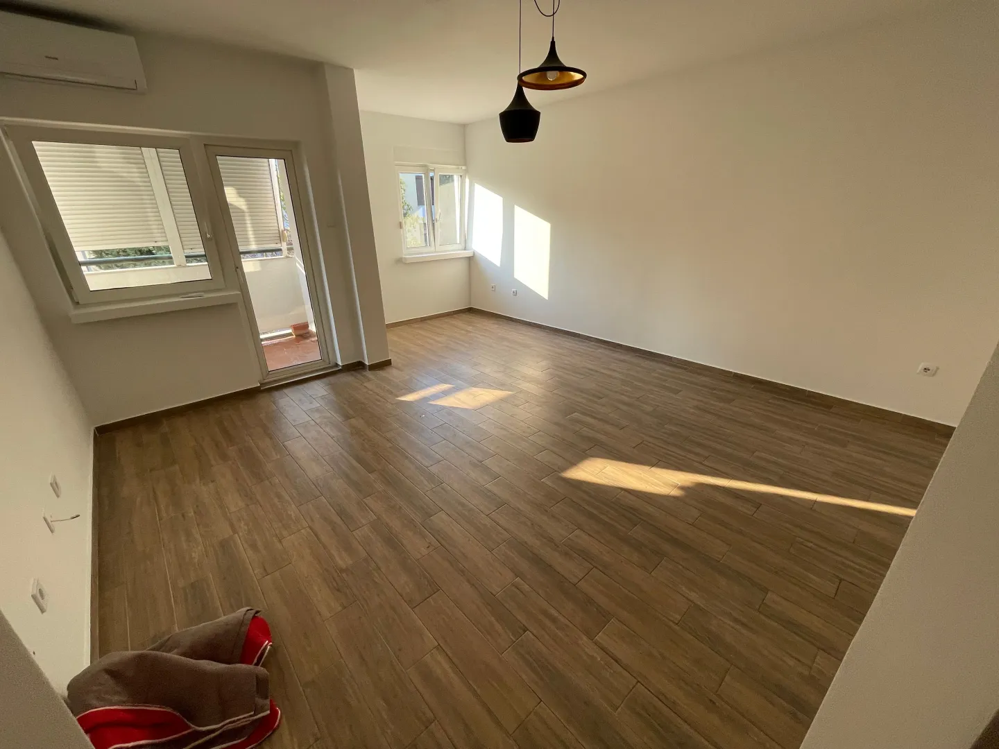 Prazna soba sa drvenim podom, bijelim zidovima i izlazom na balkon.
