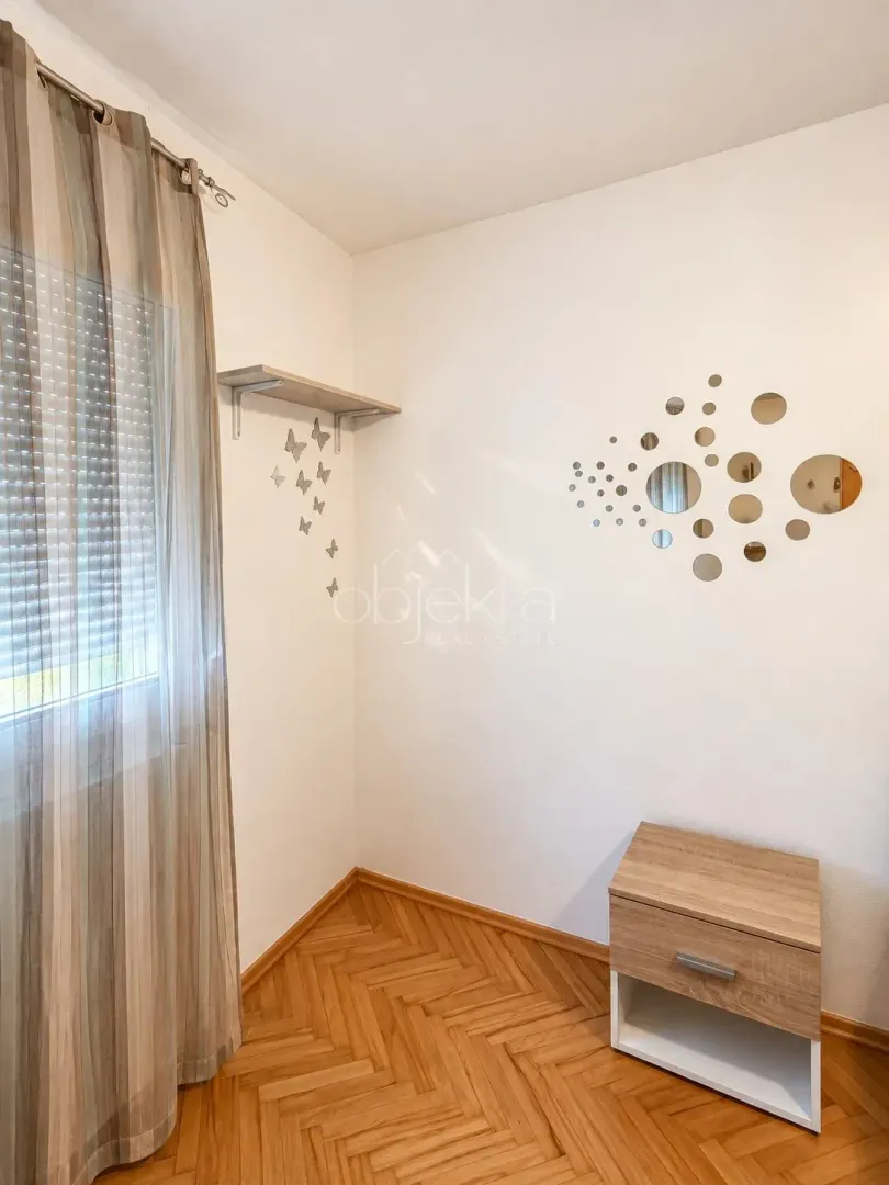 Part of a room with a wooden floor, a window with a curtain, and a small bedside table.
