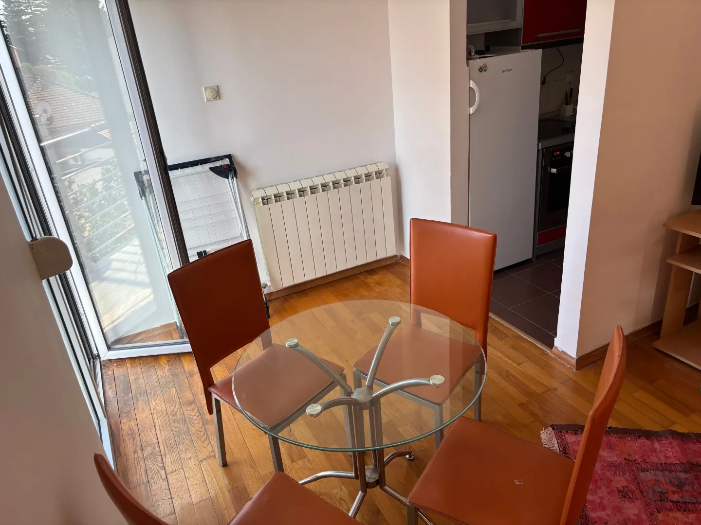 Trpezarija sa okruglim staklenim stolom i narandžastim stolicama pored izlaza na terasu.