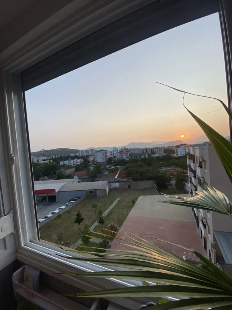 Pogled kroz prozor na grad i okolna brda pri zalasku sunca.