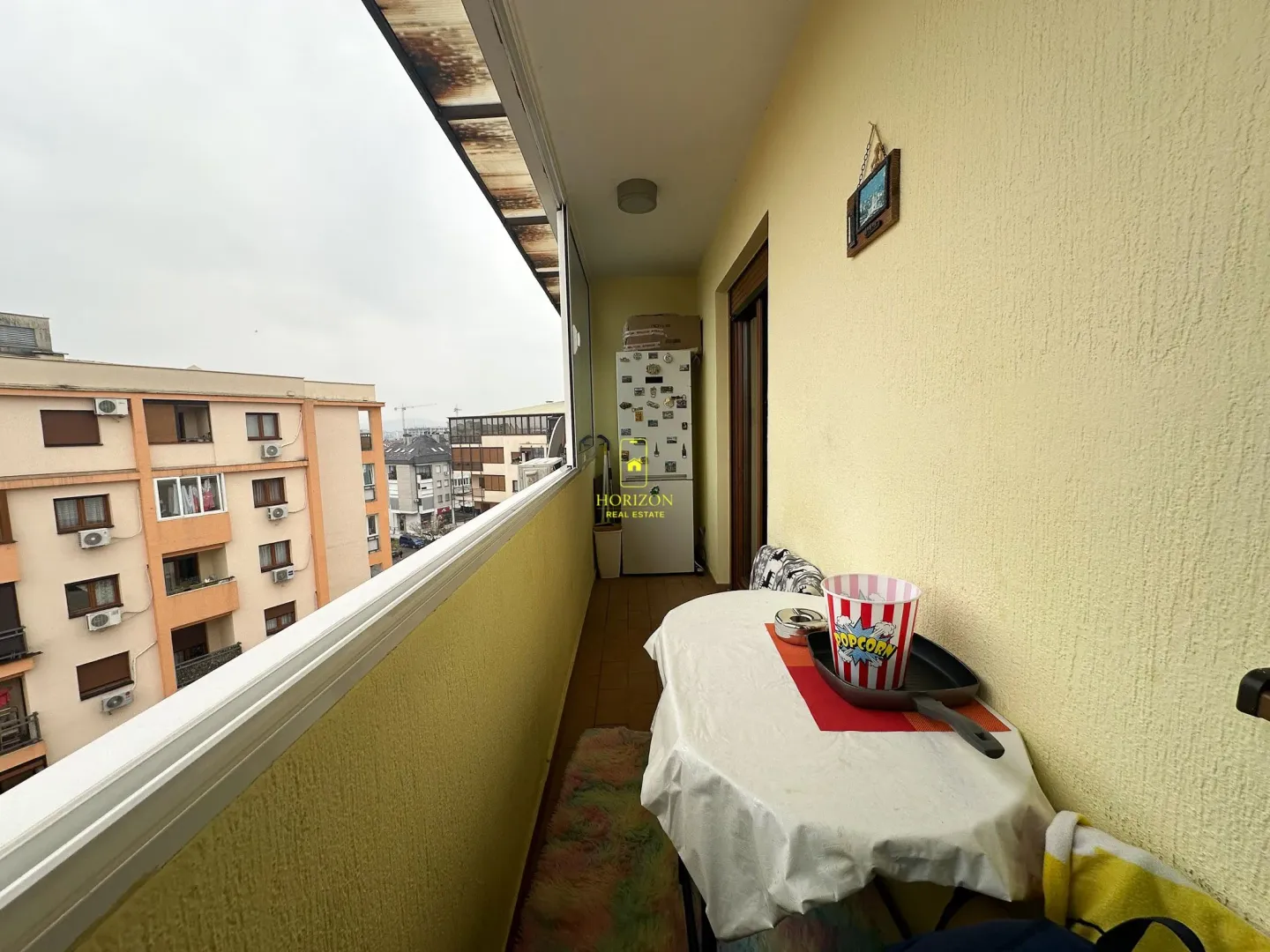 Zastakljeni balkon sa stolom i pogledom na okolne zgrade.
