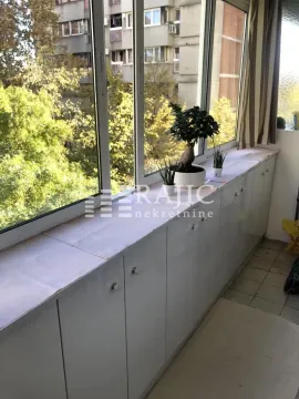 Zastakljena terasa sa ugrađenim ormarićima i pogledom na okolne zgrade. - image 10