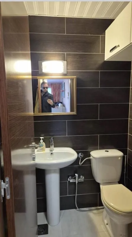 Manje kupatilo sa tamnim zidnim pločicama, umivaonikom i wc šoljom.