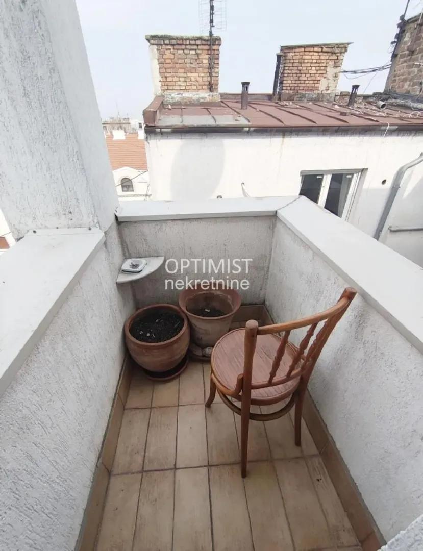 Manji popločani balkon sa drvenom stolicom i saksijama sa biljkama.