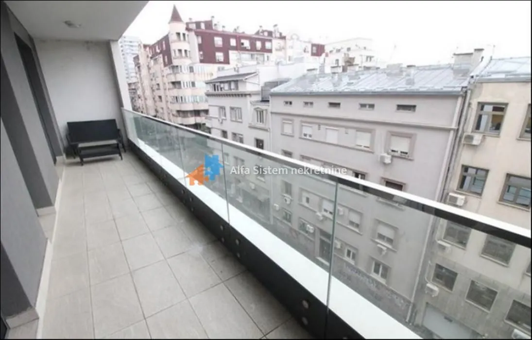 Dugačak popločani balkon sa staklenom ogradom i pogledom na okolne zgrade.