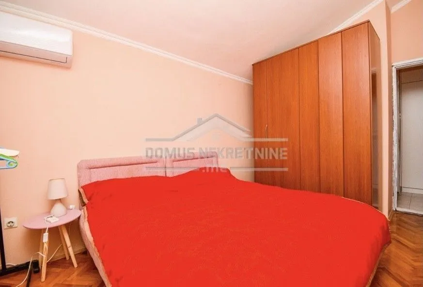 A simple bedroom with a double bed, a wooden wardrobe, and an air conditioning unit.