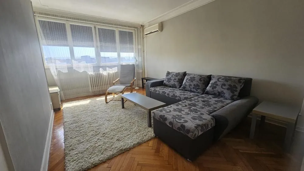 Dnevna soba sa ugaonom garniturom, tepihom i velikim prozorom.