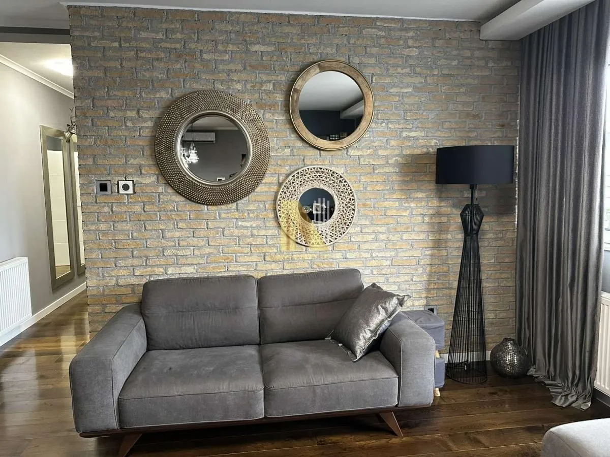 Uređena dnevna soba sa sivom garniturom, dekorativnom ciglom na zidu i ogledalima.