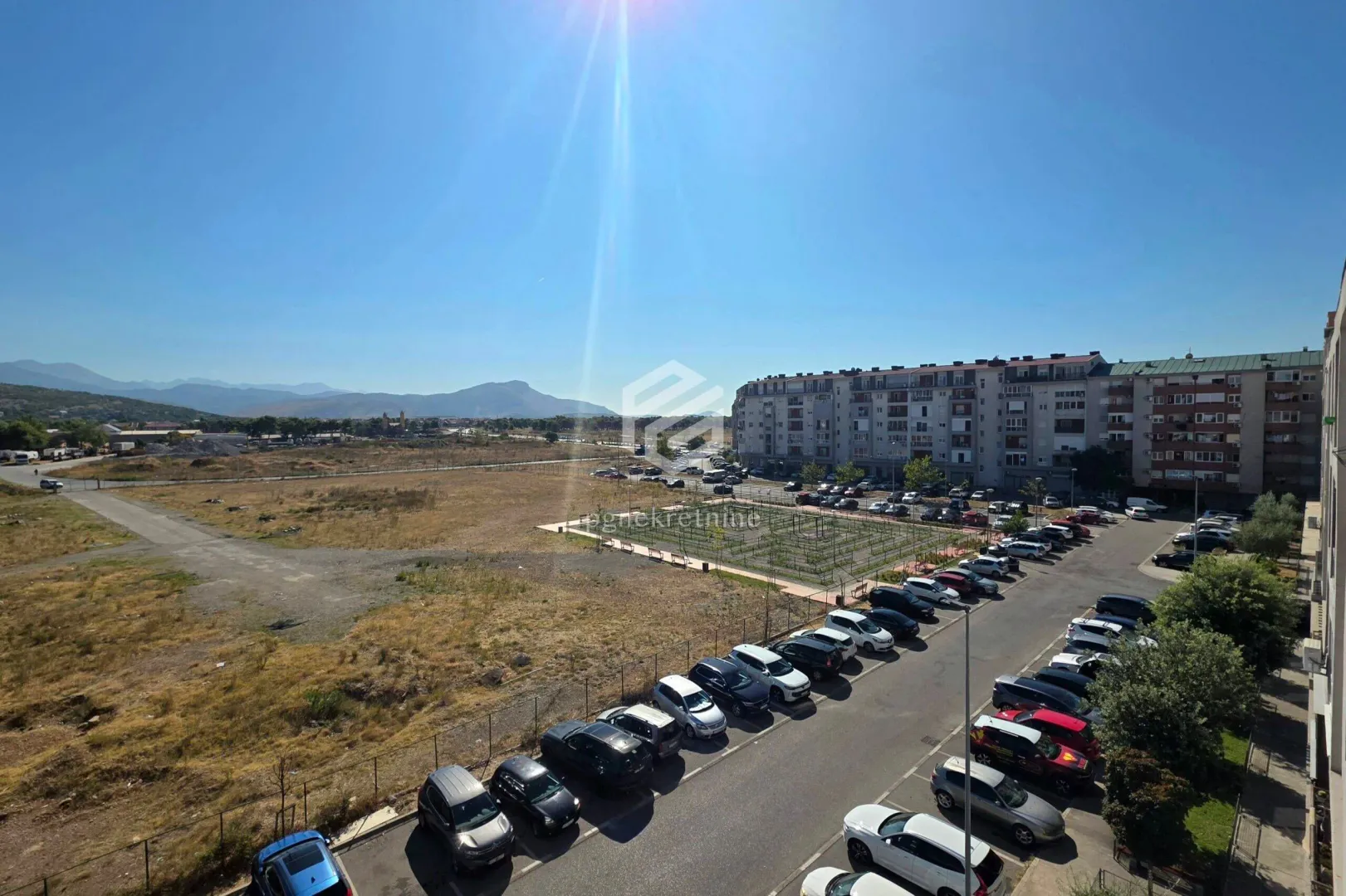 Pogled sa terase na parking prostor, okolne zgrade i planine u daljini.
