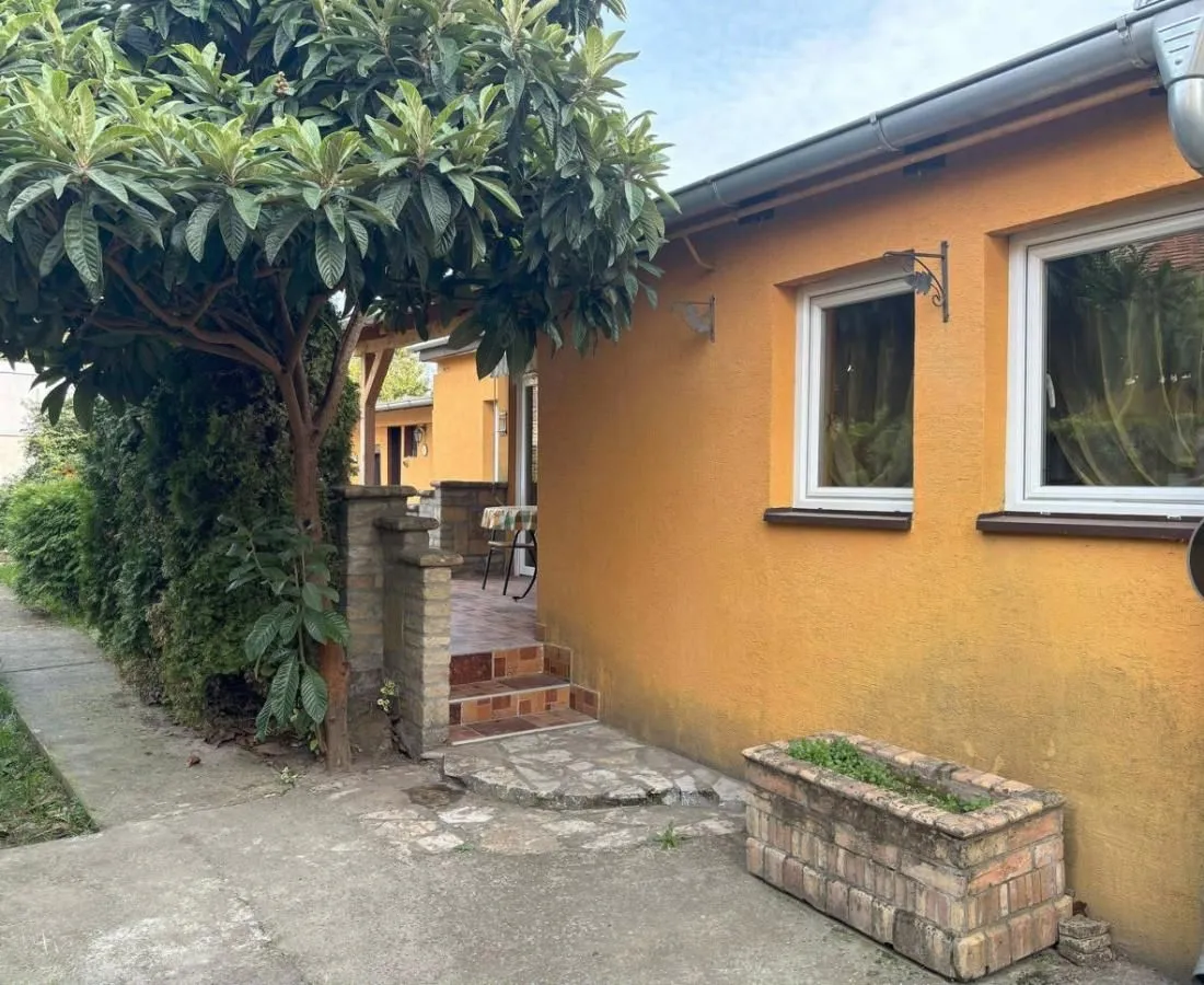 Pogled na žutu fasadu kuće sa dvorištem i zelenilom.