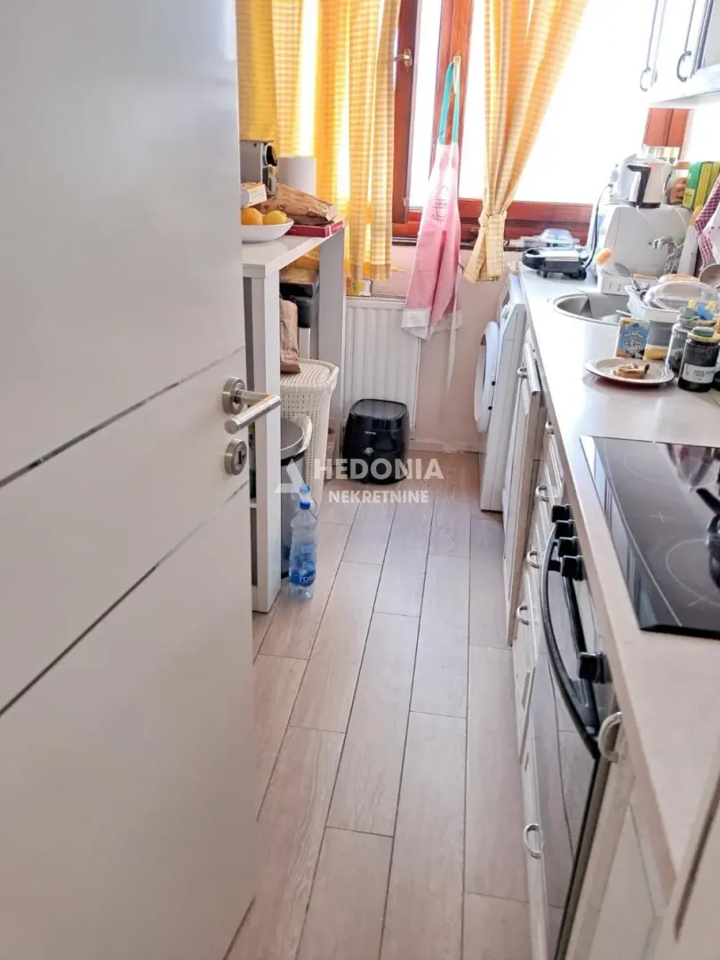 Pogled na manju kuhinju sa ugradnom pločom, sudoperom i prozorom sa žutim zavesama.