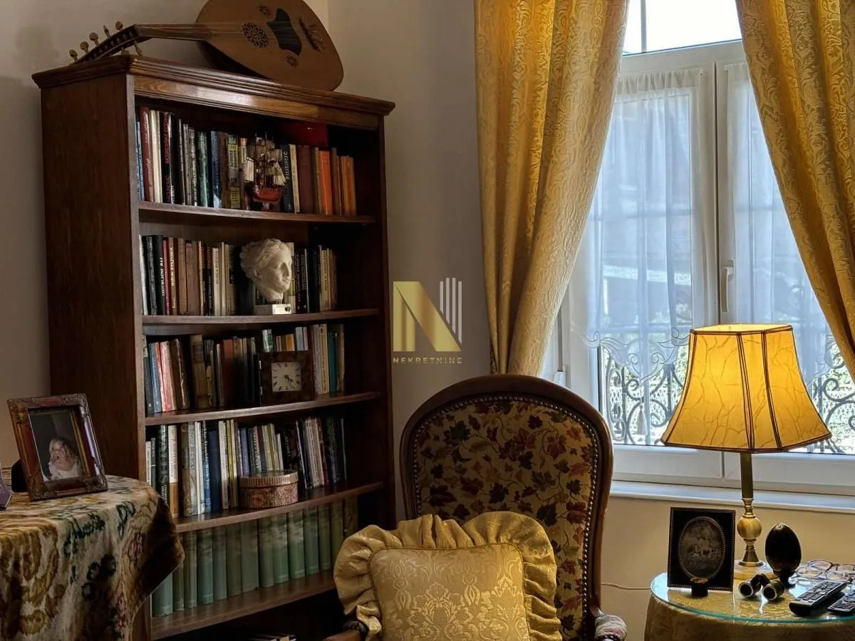 Dnevna soba sa drvenom bibliotekom, foteljom i prozorom sa žutim zavesama.