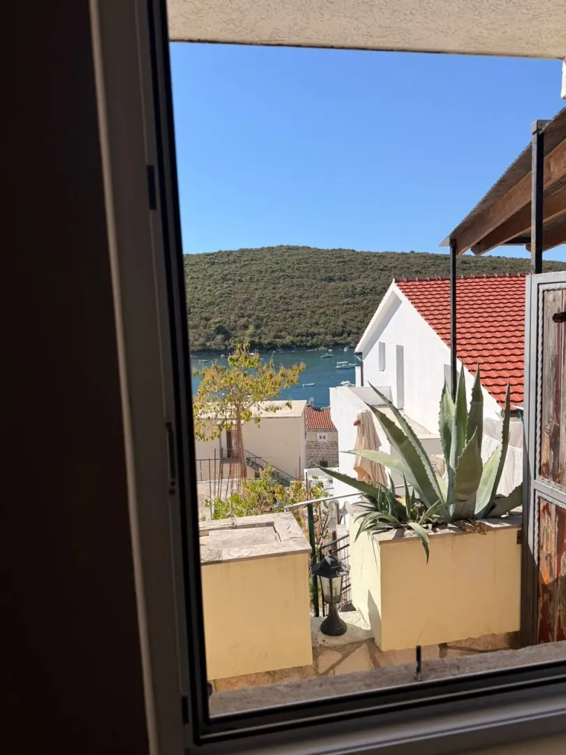 Pogled kroz prozor na mediteranske kuće, more i brda.
