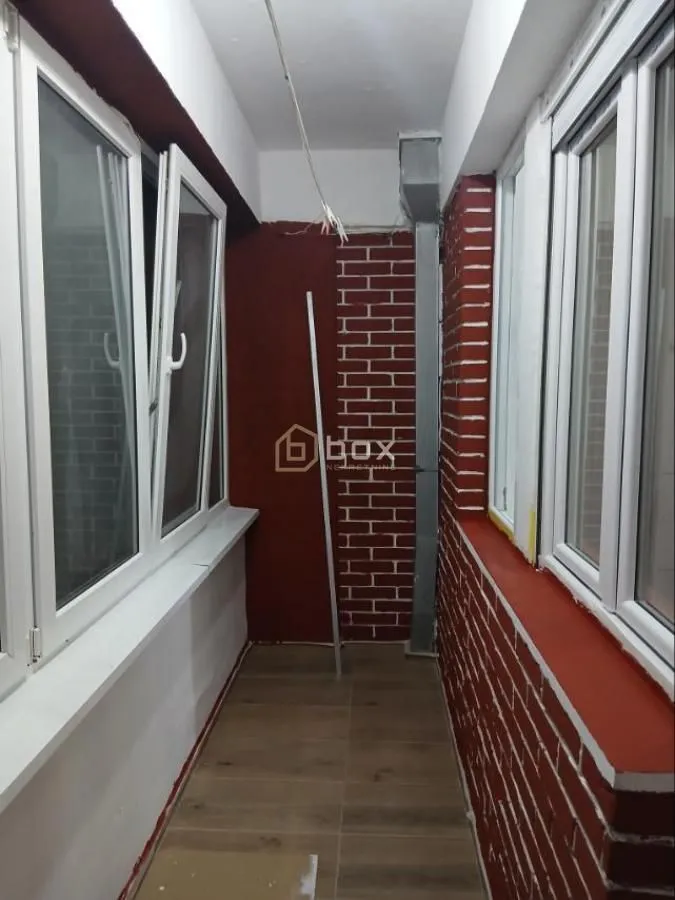 Zastakljeni balkon sa crvenim zidom od opeke i keramičkim pločicama na podu.