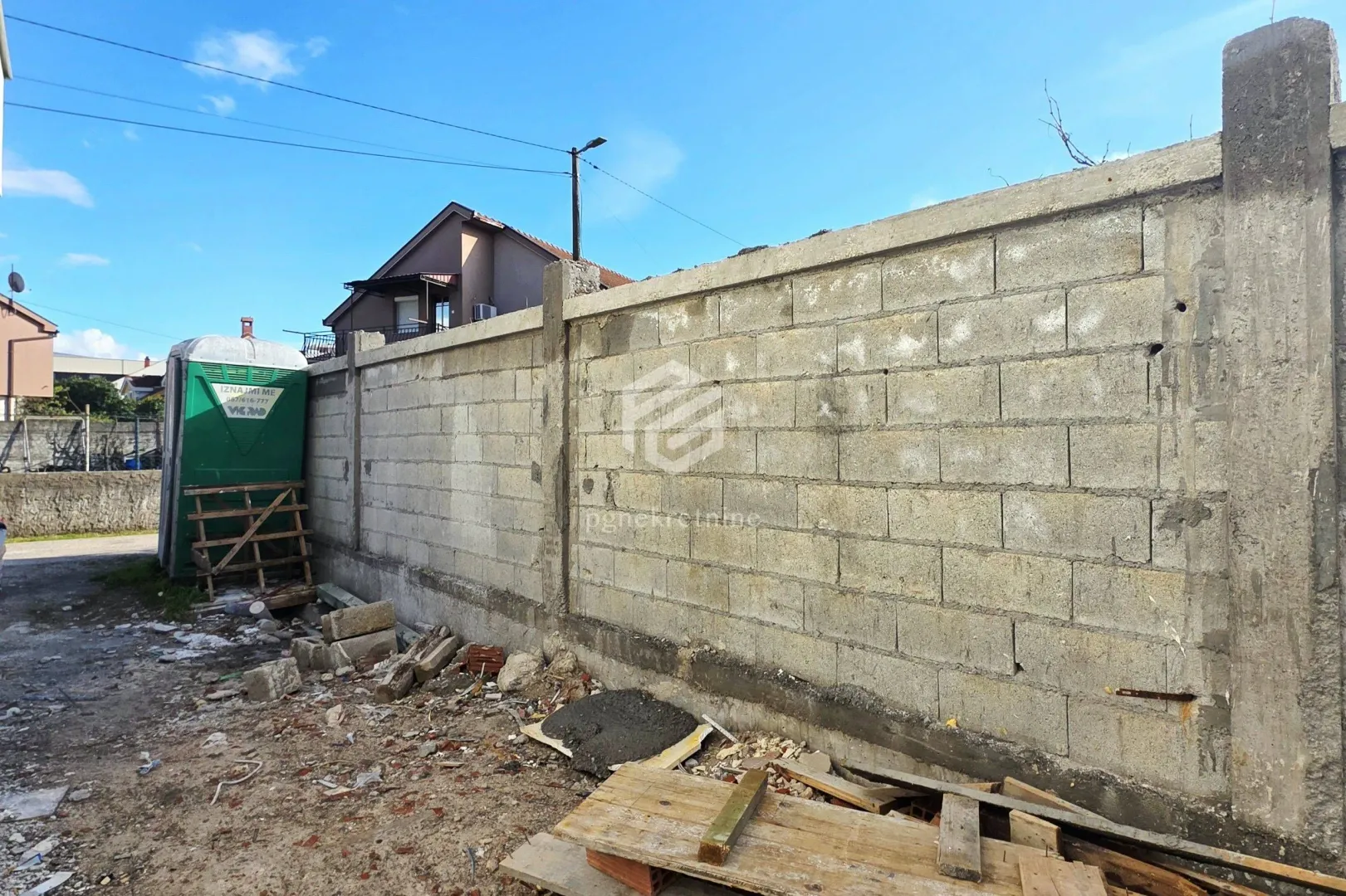 Betonski zid i dvorište sa građevinskim materijalom i pokretnim toaletom.