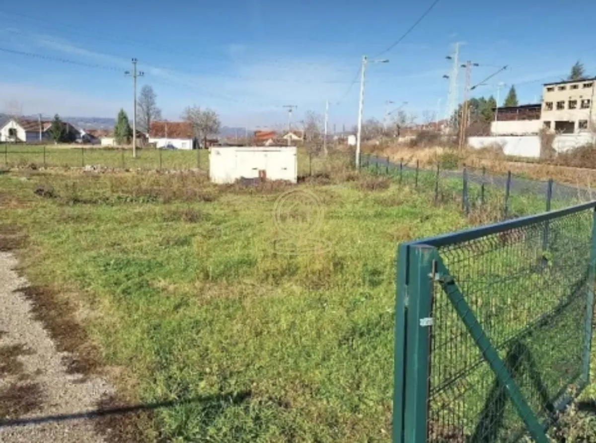 Pogled na ograđeni plac sa travnatom površinom i okolnim objektima.