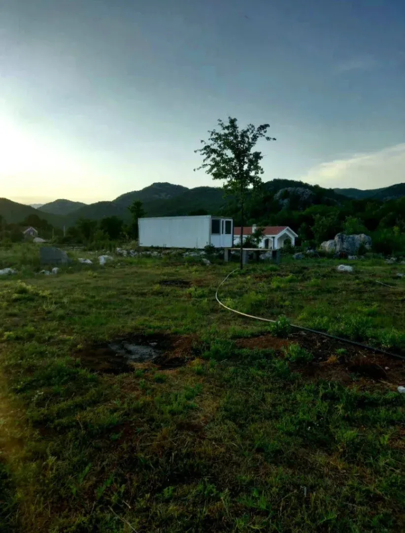 Pogled na plac sa manjim montažnim objektom i brdima u pozadini.