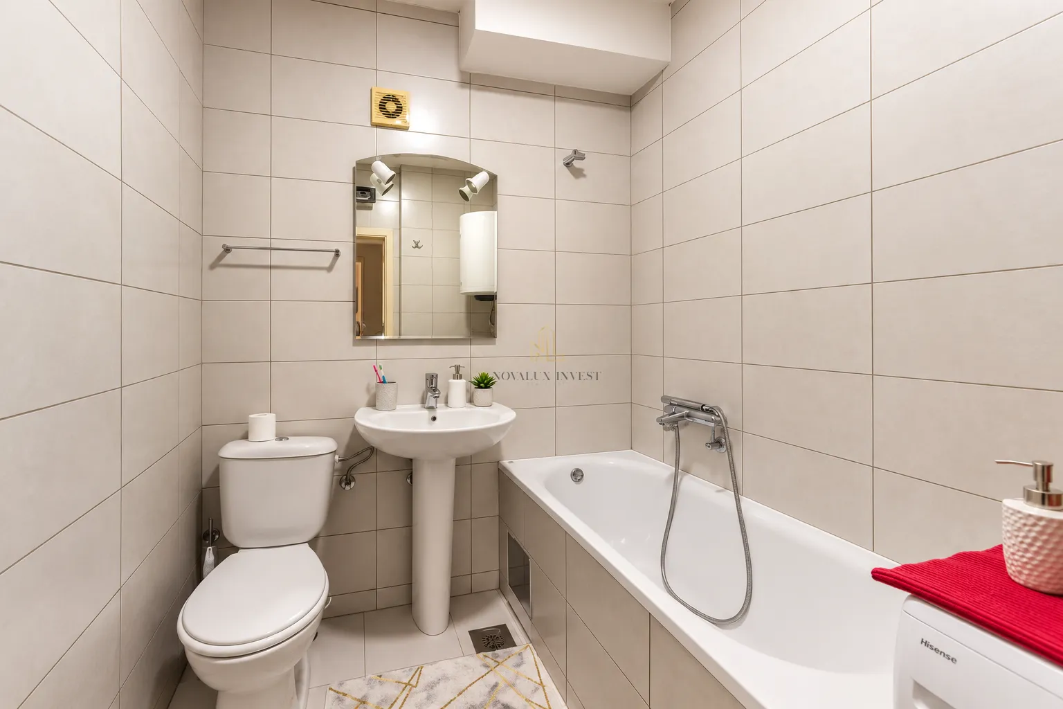 Kompaktno kupatilo sa kadom, umivaonikom i wc šoljom obloženo svijetlim pločicama.