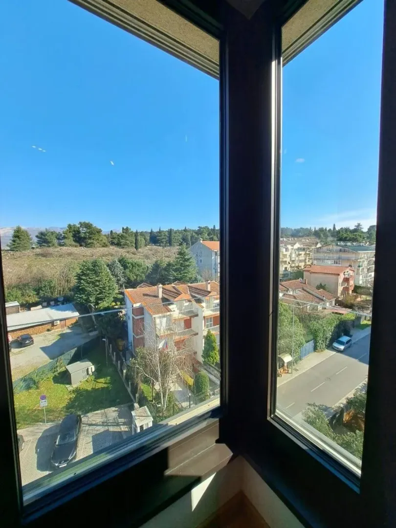 Pogled iz prozora na okolne zgrade i zelene površine.