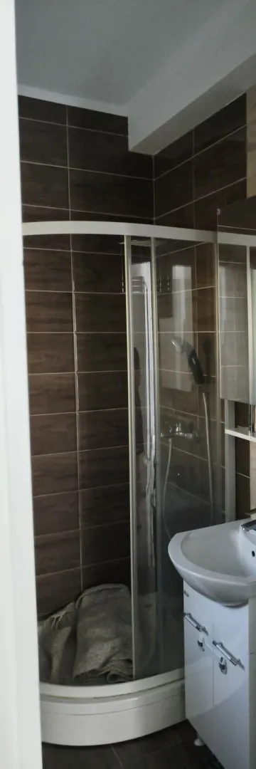 Manje kupatilo sa tuš kabinom i umivaonikom obloženo tamnim pločicama.