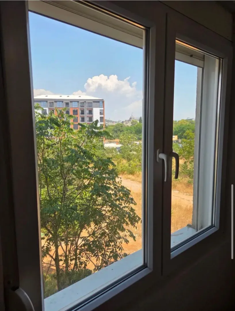 Pogled kroz prozor na zelene površine i susjedne zgrade.