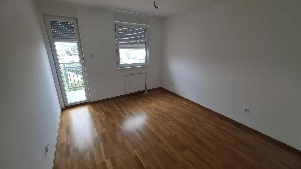 Prazna soba sa parketom, prozorom i balkonskim vratima.