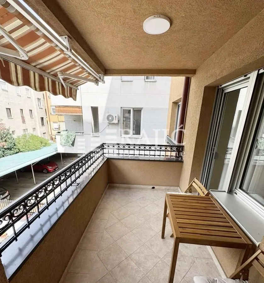 Natkrivena terasa sa drvenim stolom i pogledom na okolne zgrade.