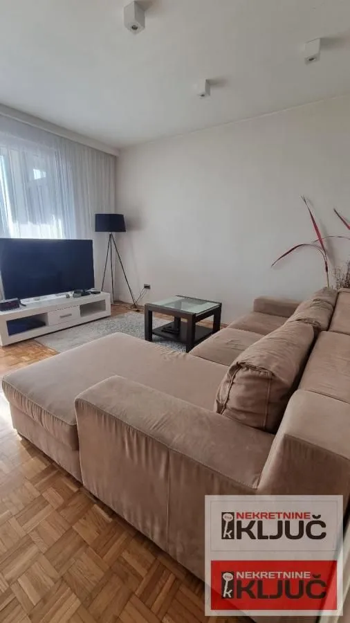 Dnevna soba sa ugaonom garniturom, televizorom i parketom na podu.